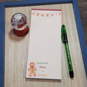 May include: A white notepad with a gingerbread man illustration and the text "EVERYONE loves a ginger" on a wooden surface with a snow globe and a green pen.