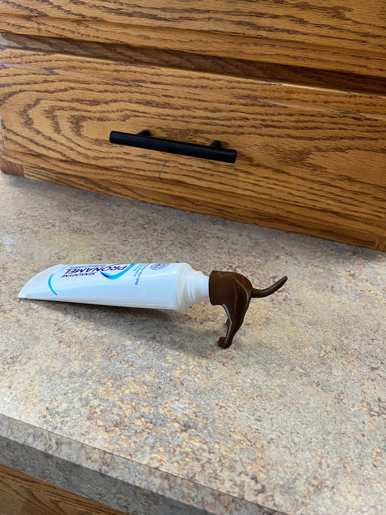 May include: A white tube of ProNamel toothpaste with a brown plastic dog-shaped dispenser. The dispenser is shaped like a dachshund and has a long tail.
