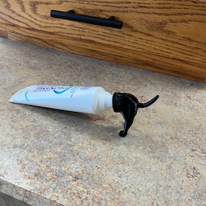 May include: A white tube of toothpaste with a black dog-shaped squeeze dispenser. The dispenser is shaped like a dog standing on its hind legs with its tail curled up.