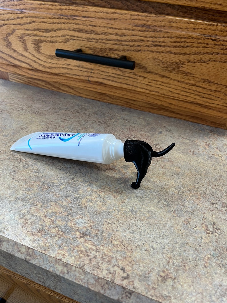 May include: A white tube of toothpaste with a black dog-shaped squeeze dispenser. The dispenser is shaped like a dog standing on its hind legs with its tail curled up.