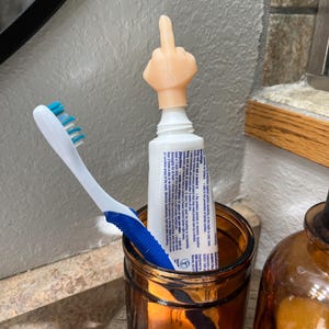 May include: A brown glass jar with a white toothbrush and a tube of toothpaste with a middle finger shaped topper. The toothpaste tube has a white label with black text.