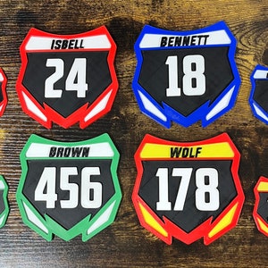 May include: Six different motocross number plates in various colors and sizes. The plates feature black backgrounds with white numbers and text. The plates are made of plastic and have a raised edge around the perimeter. The text on the plates includes "ISBELL 24", "BENNETT 18", "BROWN 456", and "WOLF 178".