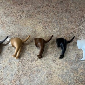 May include: Five 3D printed dog bottom hooks in different colours: black, brown, gold, white, and grey. The hooks are shaped like a dog's hind legs and tail, with the tail curled up to form a hook.