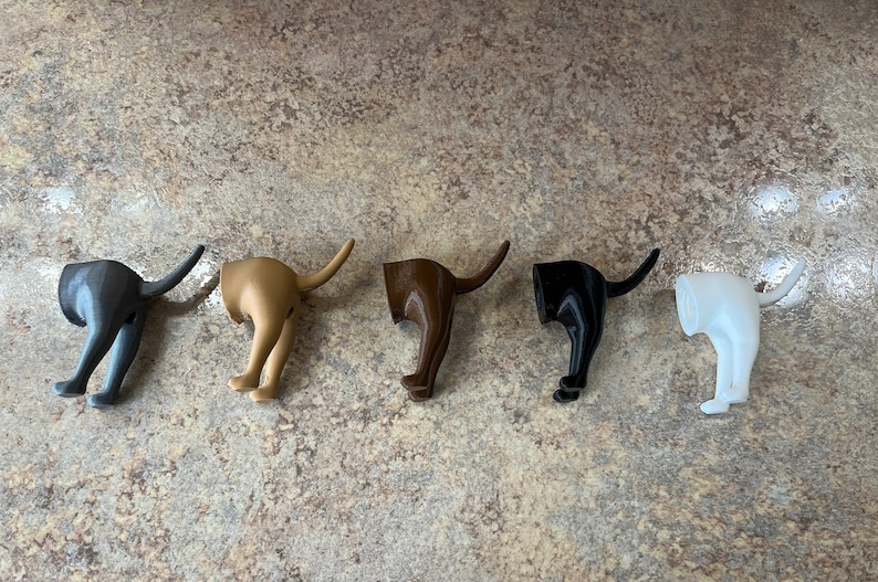 May include: Five 3D printed dog bottom hooks in different colours: black, brown, gold, white, and grey. The hooks are shaped like a dog's hind legs and tail, with the tail curled up to form a hook.