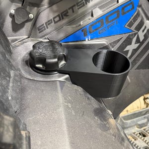 May include: Black 3D-printed cup holder attached to a Polaris Sportsman 1000 EFI ATV. The cup holder is mounted near the fuel cap. The ATV is gray and black with blue accents. The text "SPORTSMAN 1000 EFI" is visible.