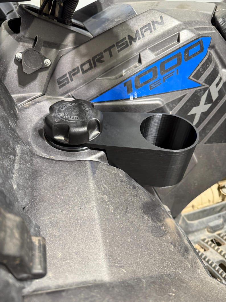 May include: Black 3D-printed cup holder attached to a Polaris Sportsman 1000 EFI ATV. The cup holder is mounted near the fuel cap. The ATV is gray and black with blue accents. The text "SPORTSMAN 1000 EFI" is visible.