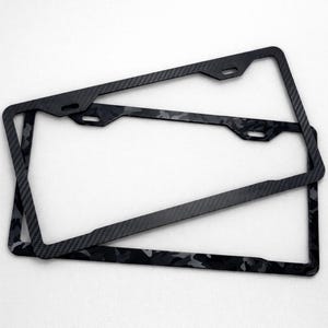 May include: Three black license plate frames with a carbon fiber and camouflage pattern. The frames are rectangular with rounded corners and have mounting holes at the top. The camouflage pattern is a mix of gray and black.