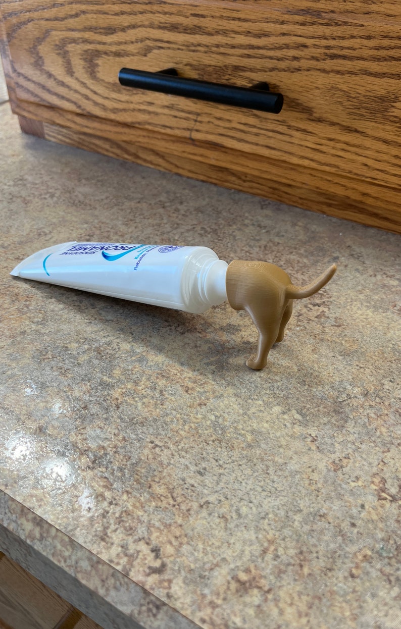 May include: A white tube of toothpaste with a brown dog-shaped squeeze dispenser. The dispenser is shaped like a dog's hindquarters and tail.