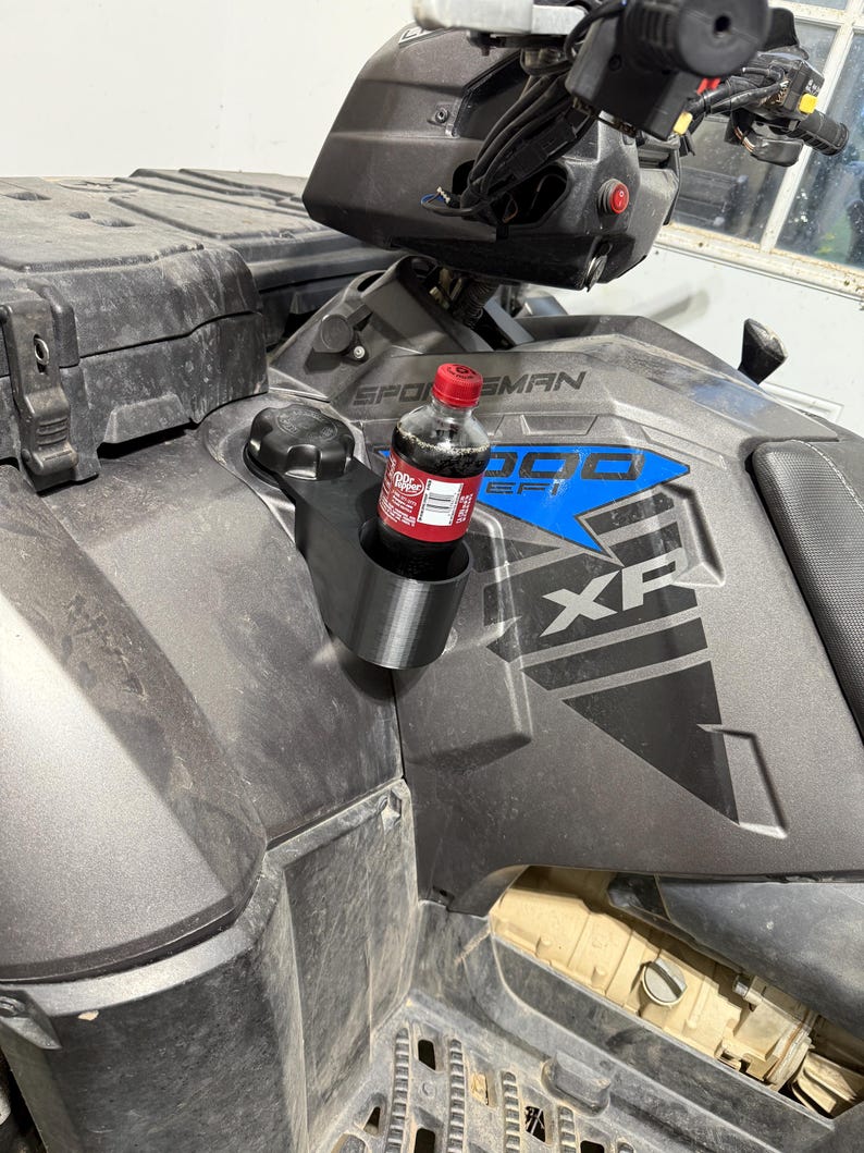 May include: A black 3D-printed cup holder attached to a gray ATV. A small bottle of Dr. Pepper is in the cup holder. The ATV has the words "SPORTSMAN 1000 XP" in blue and white.