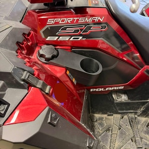 May include: A close-up of a red Polaris Sportsman SP 850 HD ATV. The ATV has a black cup holder and a black fuel cap. The ATV is parked on a dirty surface.