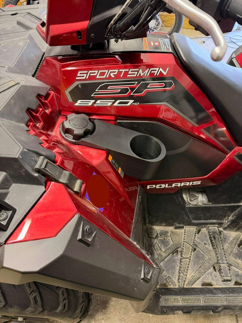 May include: A close-up of a red Polaris Sportsman SP 850 HD ATV. The ATV has a black cup holder and a black fuel cap. The ATV is parked on a dirty surface.