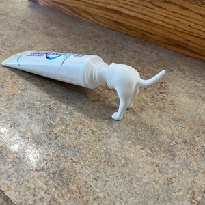May include: A white plastic dog-shaped toothpaste dispenser attached to a tube of toothpaste.