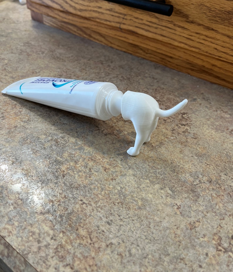 May include: A white plastic dog-shaped toothpaste dispenser attached to a tube of toothpaste.