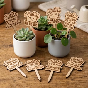 Può includere: Paletti di legno per piante con disegni di vasi incisi e frasi umoristiche come "Ti ho dato fiducia!" e "Perché sono così?". I paletti sono esposti in piccole piante in vaso, su una superficie di legno.