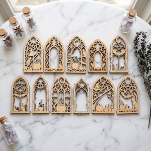 May include: A collection of laser-cut wooden gothic window ornaments. Each ornament features intricate designs with witchy and Halloween themes, including cauldrons, bats, and cats. The ornaments are arranged on a marble surface.