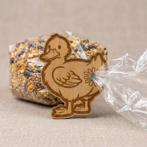May include: A wooden duck-shaped bag clip, with a detailed outline of a duck with a smiling face and webbed feet. The clip is holding a clear plastic bag, which is attached to a bag of birdseed. The clip is made of light-coloured wood.