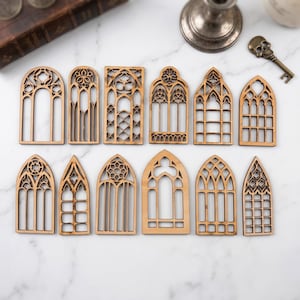 May include: A collection of twelve laser-cut wooden gothic window shapes in various designs. The windows are light brown and feature intricate tracery patterns, perfect for crafting or decorative projects. The windows are arranged on a white marble surface.