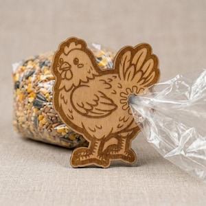 May include: A wooden chicken-shaped bag clip. The clip is light brown with etched details, holding a clear plastic bag. The bag contains birdseed. The clip is designed to seal bags and keep contents fresh.