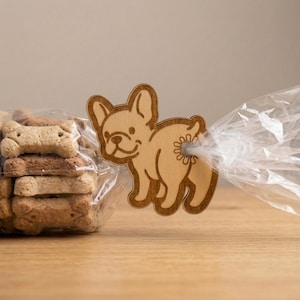 May include: A wooden dog-shaped bag clip, featuring a French bulldog design, is attached to a clear plastic bag. The bag contains dog biscuits. The clip is made of light-colored wood and has a detailed engraved design.