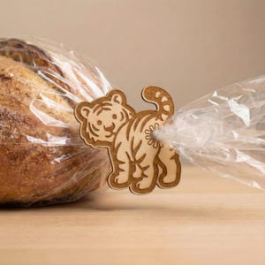 May include: A wooden tiger-shaped bread clip, with etched details, secures a clear plastic bag around a loaf of bread. The clip is light brown and features a smiling tiger design. The bread is a golden brown color.