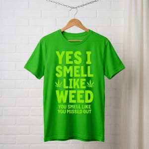 May include: A vibrant green t-shirt featuring the phrase "YES I SMELL LIKE WEED" in bold, lime green lettering. The design includes cannabis leaf symbols and the text "YOU SMELL LIKE YOU MISSED OUT". The shirt is displayed on a wooden hanger.