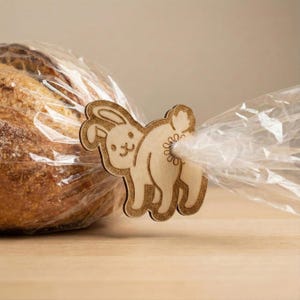 Puede incluir: Un clip de pan con forma de conejito de madera, con un contorno detallado, asegura una bolsa de plástico transparente alrededor de una barra de pan. El clip es de color marrón claro y presenta un lindo diseño de conejito.