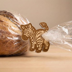May include: A wooden zebra-shaped bread clip, featuring etched stripes and details, secures a clear plastic bag. The clip is light brown and adds a decorative touch to the bread packaging. The background is a light brown surface.