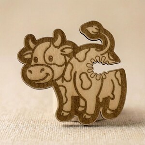 Close-up of the wooden laser-cut cow-shaped bag clip, showcasing its engraved details and the functional clip design, resting on a textured light brown fabric surface.