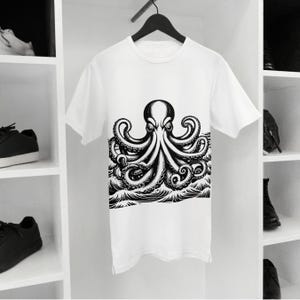 May include: White t-shirt featuring a detailed black and white illustration of an octopus. The octopus design is centered on the shirt, with its tentacles extending over stylized waves. The shirt is hanging on a black hanger.
