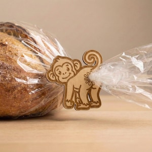 May include: A wooden monkey-shaped bread clip, with a smiling face and curly tail, secures a clear plastic bag containing a loaf of bread. The clip is made of light-colored wood and is positioned on a light brown surface.