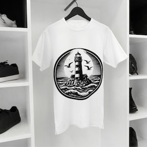 Peut inclure: T-shirt blanc avec un motif noir et blanc d'un phare sur un îlot rocheux, entouré d'oiseaux et de vagues. Le motif est dans un cercle. Le t-shirt est suspendu à un cintre noir.