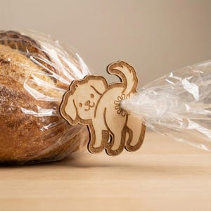 Op de afbeelding: Een houten broodzakclip in de vorm van een hond, lichtbruin van kleur. De clip houdt een doorzichtige plastic zak gesloten, met een brood in de achtergrond. De hond heeft een omhoogstaande staart en de clip heeft een speels ontwerp.