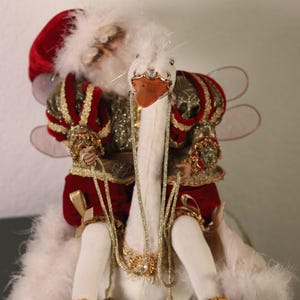 May include: A whimsical decorative figurine featuring a white swan with a jeweled beak and a rider in a red and gold outfit. The swan has a long, beaded necklace with bells. The figurine is on a gold-trimmed base.