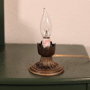 Może przedstawiać: Lampa w stylu vintage z przezroczystą żarówką w kształcie płomienia i ozdobną podstawą z brązu. Lampa stoi na ciemnozielonej powierzchni. W tle drewniany świecznik ze świecą.