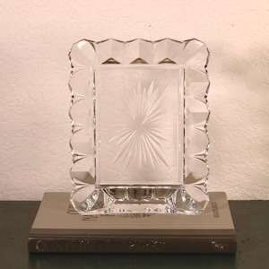 May include: A clear, rectangular glass picture frame featuring a faceted, scalloped edge and a central starburst pattern etched into the glass surface. The frame is designed to hold a photograph and stands upright on a flat, horizontal base.
