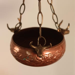 May include: A copper hanging bowl with a floral design and deer head accents. The bowl is suspended from a chain.