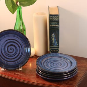 May include: Four blue ceramic plates with a spiral design. The plates are stacked on a wooden surface.