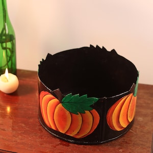 Könnte beinhalten: Ein schwarzer, runder Behälter mit einem Kürbismotiv. Die Kürbisse sind orange mit grünen Blättern. Eine grüne Glasflasche und eine brennende Kerze befinden sich im Hintergrund.
