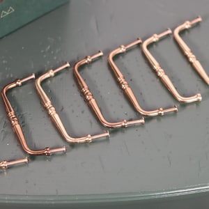 May include: Seven decorative, curved metal handles with a warm, copper-gold finish are arranged on a gray surface. Each handle has an ornate design and a small, cylindrical end. The handles are likely for furniture or decorative purposes.