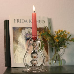 May include: A clear glass candlestick with a lit pink candle. The candlestick has a decorative design with a round base and a flared top. A book titled "FRIDA KAHLO" is in the background, along with a small vase of yellow flowers.