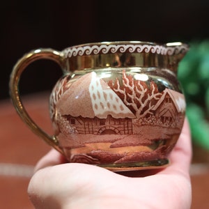 Puede incluir: Un pequeño creamer de cerámica marrón con un borde dorado y un diseño de escena de invierno blanco y marrón. El creamer tiene un asa y un pico.
