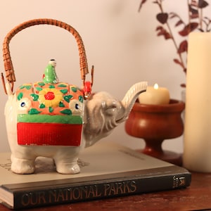 May include: A white ceramic teapot shaped like an elephant with a colorful floral design and a woven handle. The teapot is sitting on a book titled "Our National Parks".