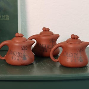 May include: Three small, reddish-brown ceramic teapots with rounded bodies, curved handles, and small lids. Each teapot has a spout and is decorated with black calligraphy and a stylized tree design.