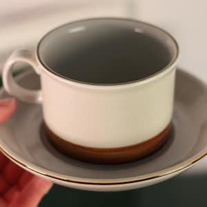 Puede incluir: Un juego de taza y platillo de cerámica blanca. La taza tiene un interior gris y una base marrón, con un platillo a juego. Ambas piezas están adornadas con una banda dorada. La taza se sostiene en una mano.