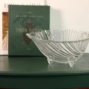 Puede incluir: Un cuenco de vidrio transparente con un diseño en espiral, apoyado sobre tres pequeños pies. El cuenco está colocado delante de dos libros, uno de ellos titulado "The Secret History Twin Peaks". Es un objeto decorativo.