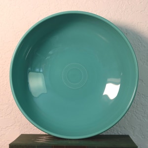 May include: A round, turquoise ceramic bowl with a smooth, glossy finish. The interior features a series of concentric circular rings embossed in the center, and the rim is slightly raised, creating a deep basin for serving or holding various items.