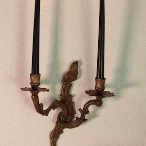 May include: An antique brass wall sconce with two lit black candles. The sconce has an elaborate, scrolling design and is mounted on a textured white wall. The candles emit a warm glow, creating a classic, elegant atmosphere.