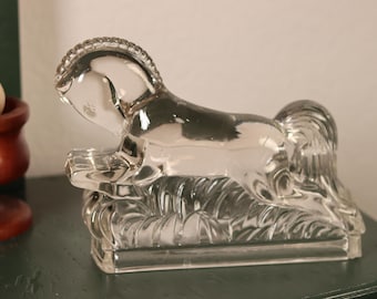 May include: A clear glass horse figurine with a detailed mane and tail, resting on a rectangular base. The horse is in a reclining position, with its head raised. The figurine is likely a decorative item.