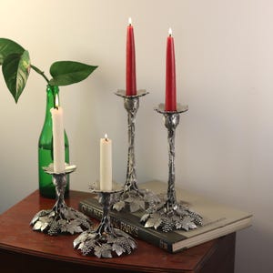 May include: Three silver candle holders with a grapevine design. Two holders have red candles and one has a white candle. The candle holders are on a wooden table with a book.