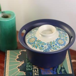 Può includere: Un piatto da portata in ceramica blu con un coperchio. Il coperchio ha uno sfondo bianco con un motivo floreale in blu, verde e oro. Il piatto è appoggiato su un libro.